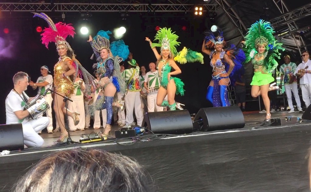 5 ladies dressed in bright colourful bodysuits or skirt and top combos, and hats with large colourful feathers, dancing on stage together at the Newham Show.