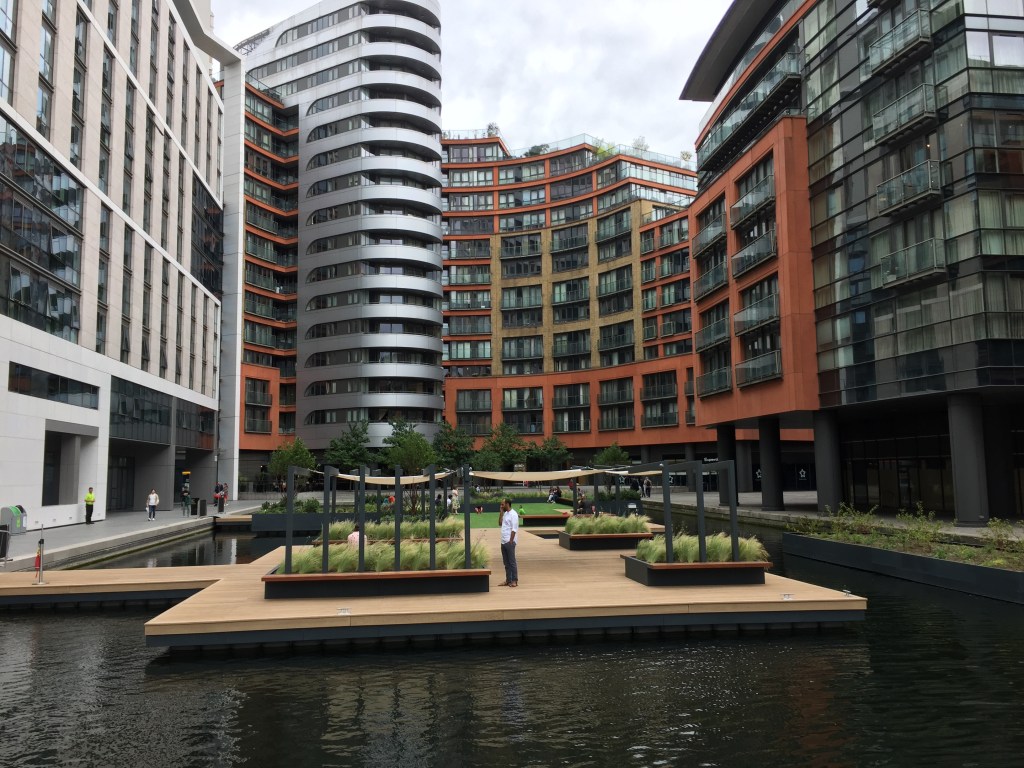 The Floating Pocket Park in Merchant Square. This is a small area divided equally into 2 sections of paving and grass, and is connected to the shore of the canal by narrow pathways. The park contains beds of greenery, with low walls around them just wide enough for people to sit on.