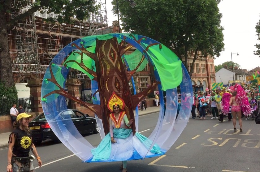A lady at the Newham Carnival wearing a light blue dress, the skirt of which connects at the bottom to a large, tall sphere that encircles her, and has an artistic representation of a tree with a brown trunk and green leaves behind her.