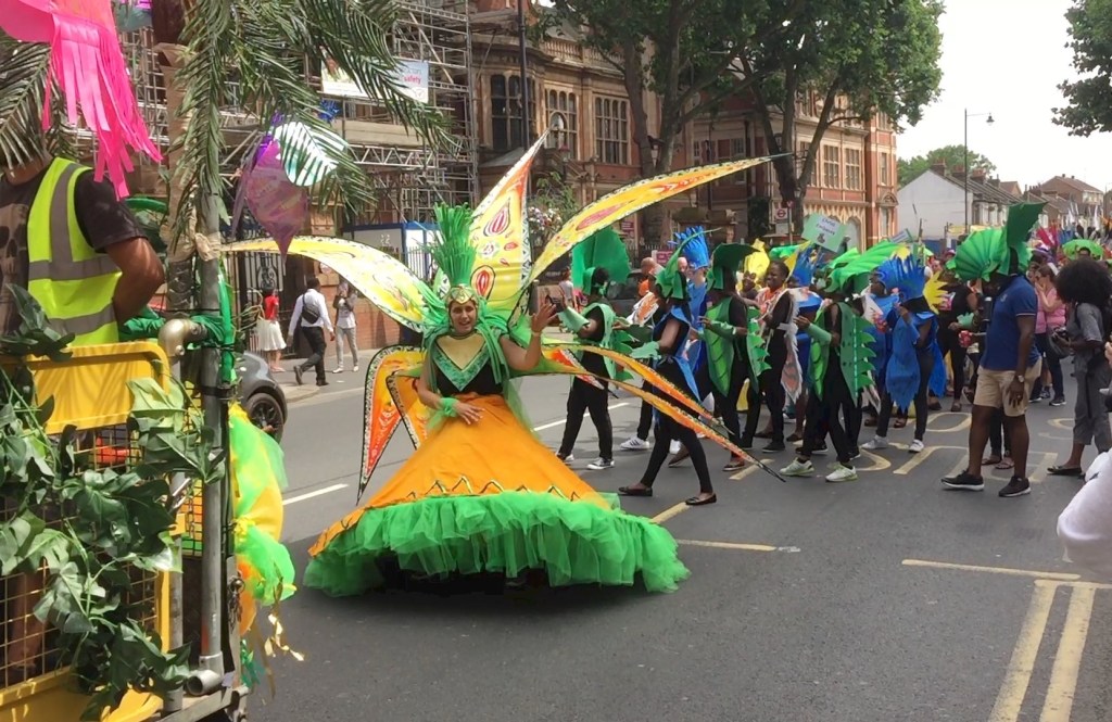 A lady in the Newham Carnival wearing a dress that has a wide orange skirt with a big leafy green hem, and a hat with tall green leaves on it. Attached to her back are some very large yellow leaves with patterns on them that are spread out behind her.