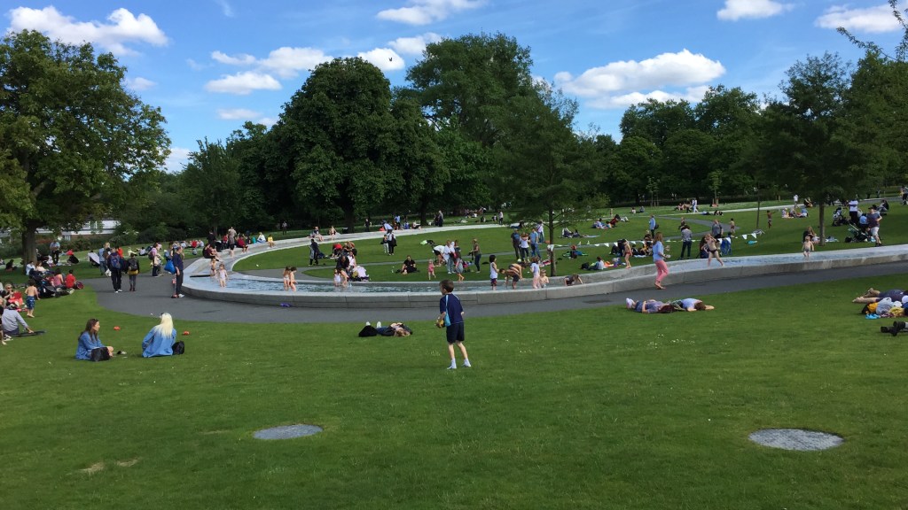 Children playing in the Princess Diana Memorial Fountain in Hyde Park. The fountain is a long, narrow channel of flowing water that encircles a large area of grass in Hyde Park.