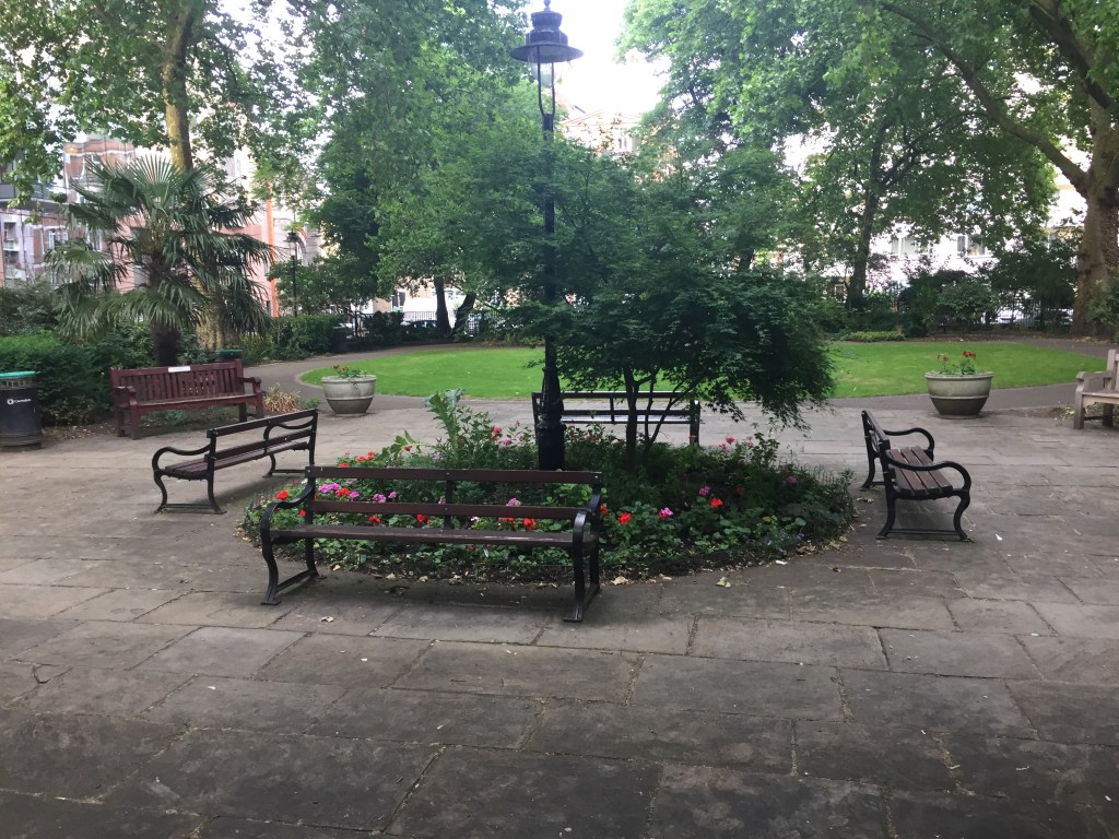 Benches placed around a circular flower bed, which also has a few very small trees and a tall lamppost in the centre, in Red Lion Square.