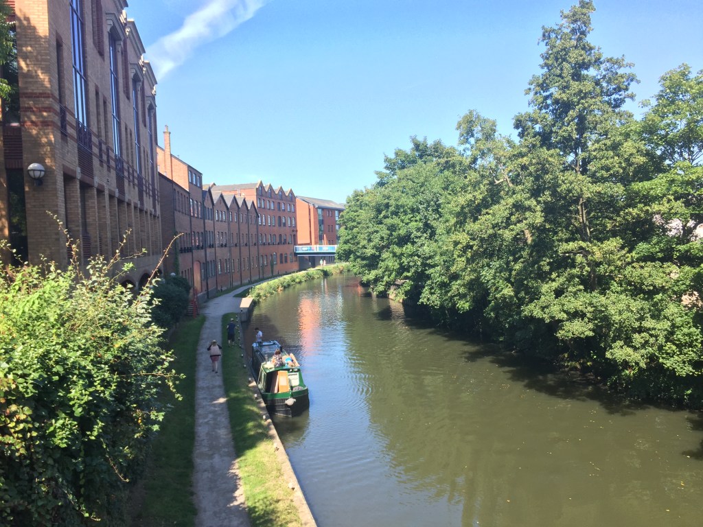The River Wey in Guildford. A green canal boat is moored on the left, next to a footpath running alongside the river, passing tall buildings of 3 and 4 storeys. The right side of the river is lined with leafy trees that hang low over the water.