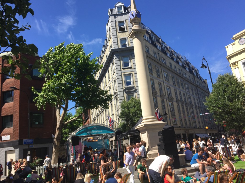 A big crowd of people walking and sitting around the tall pillar structure in the centre of the Seven Dials district, a large circular space where 7 shopping streets converge.