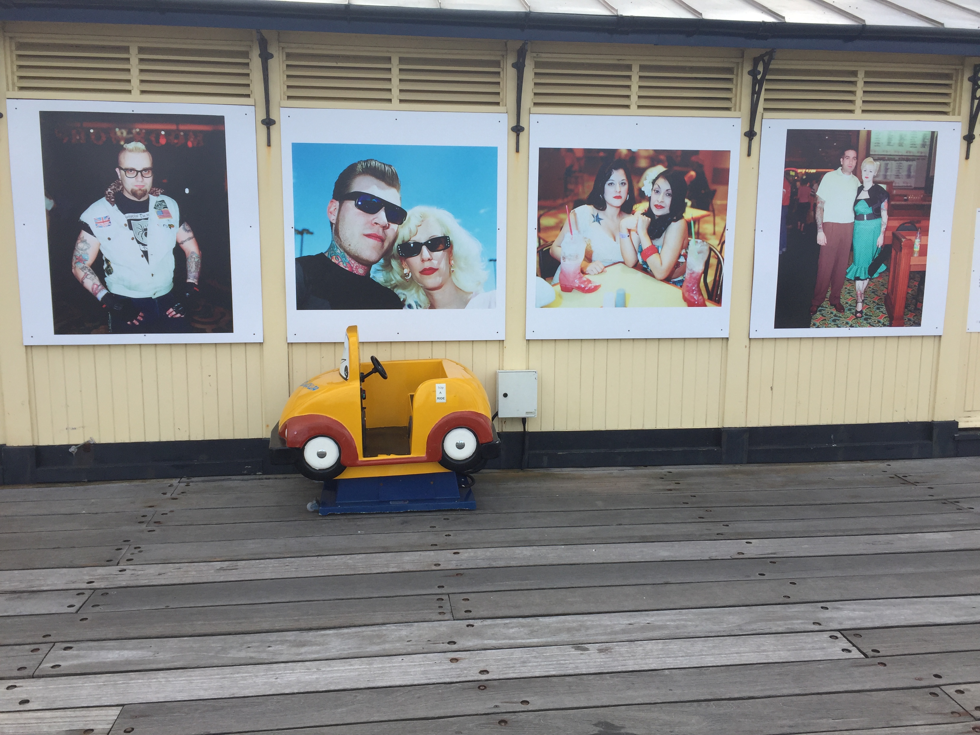 A wall in a shelter on Southend Pier, with photo posters of different people - a man with a goatee, a man and a woman posing while wearing sunglasses, 2 ladies having drinks together, and a man and woman standing together.