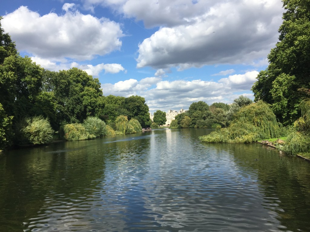 Large lake in St James's Park, with lots of green buses and trees on each side, and a big building at the end of the lake in the far distance.