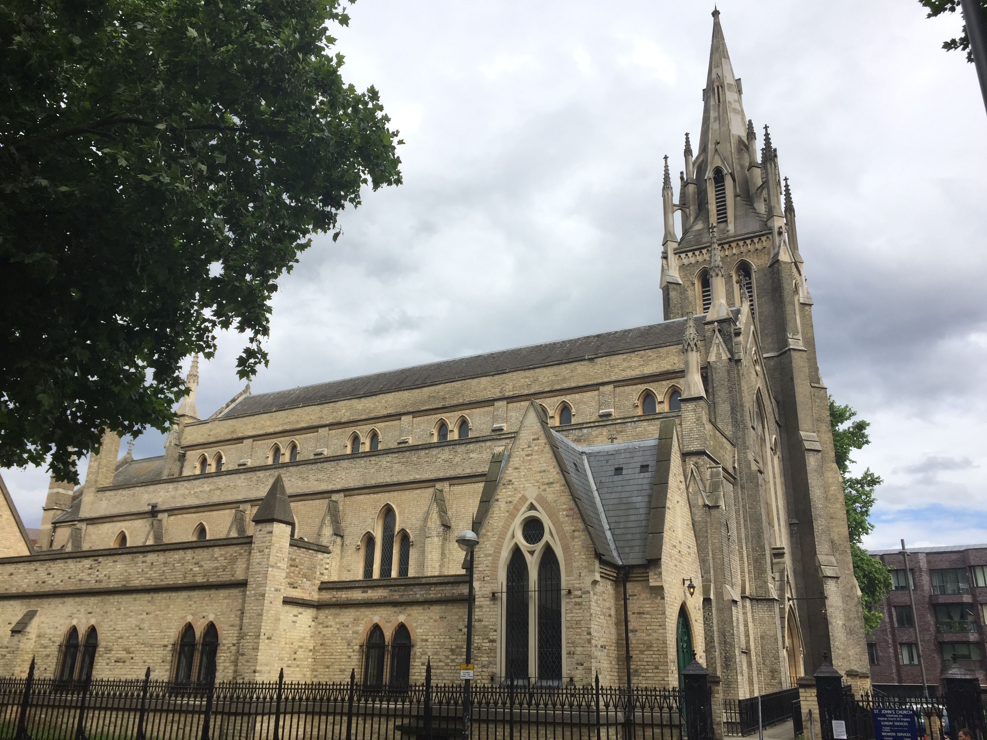 St John's Church in Stratford, a long building with each of the 3 floors set back slightly from the one beneath it, like steps on a staircase. There is a tall ornate spire at the end of the building.
