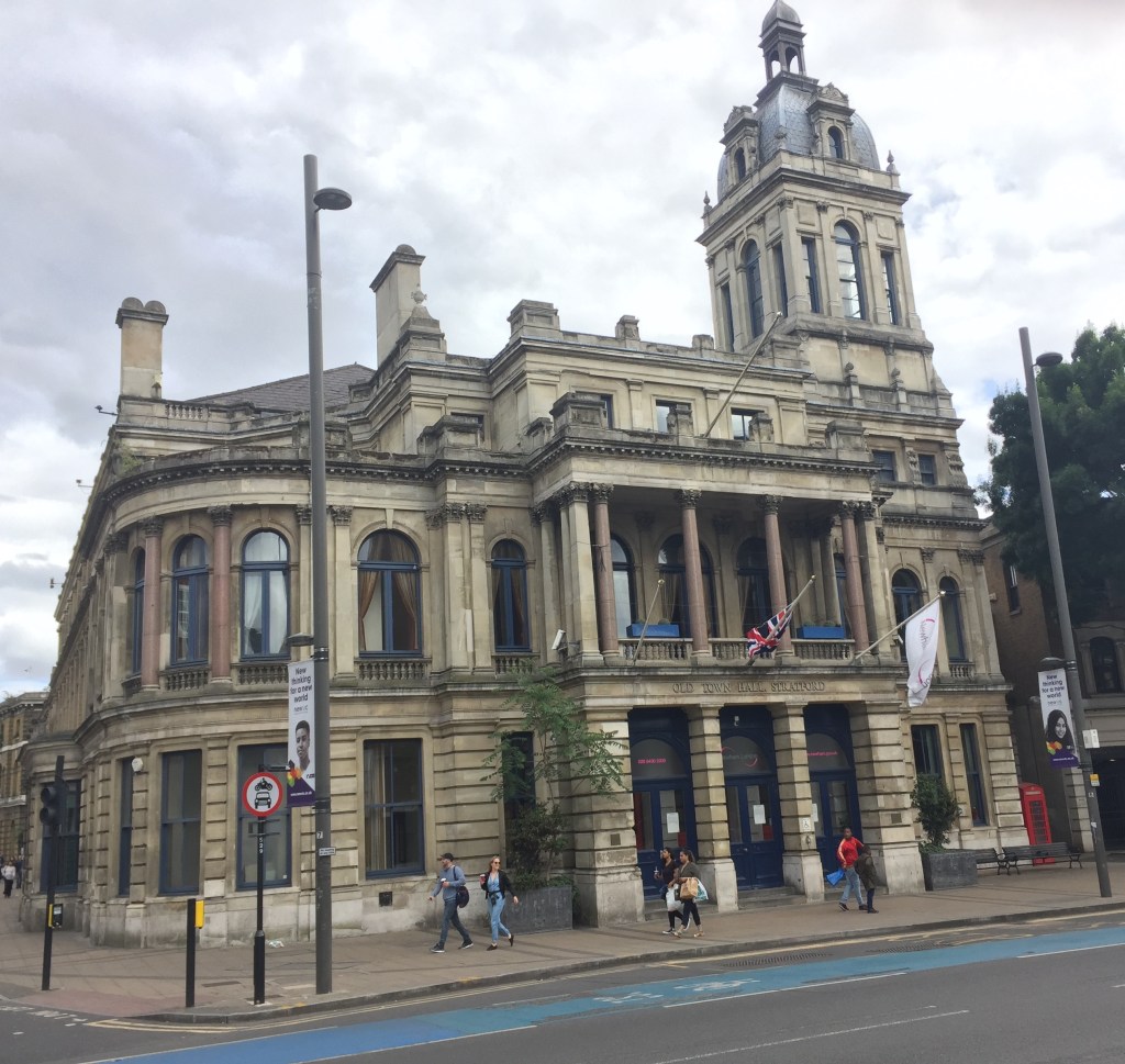 The Old Town Hall in Stratford, a 3-storey stone building with pillars in front of the ground floor entrance and the first floor balcony. A Union Jack and a white flag with the Newham London logo are hanging on poles sticking out from the balcony. The right side of the building extends up to an ornate tower.