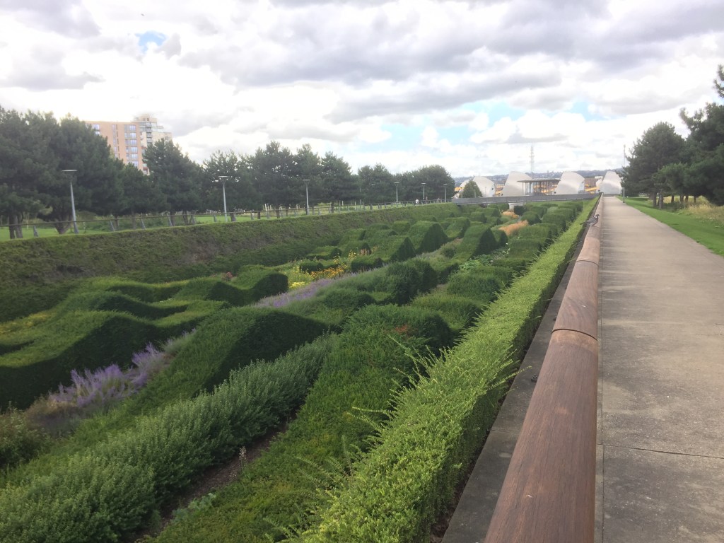 Long lines of hedges that curve up and down randomly like undulating waves, with a few colourful flowers in between them, leading towards the Thames Barrier in the distance.