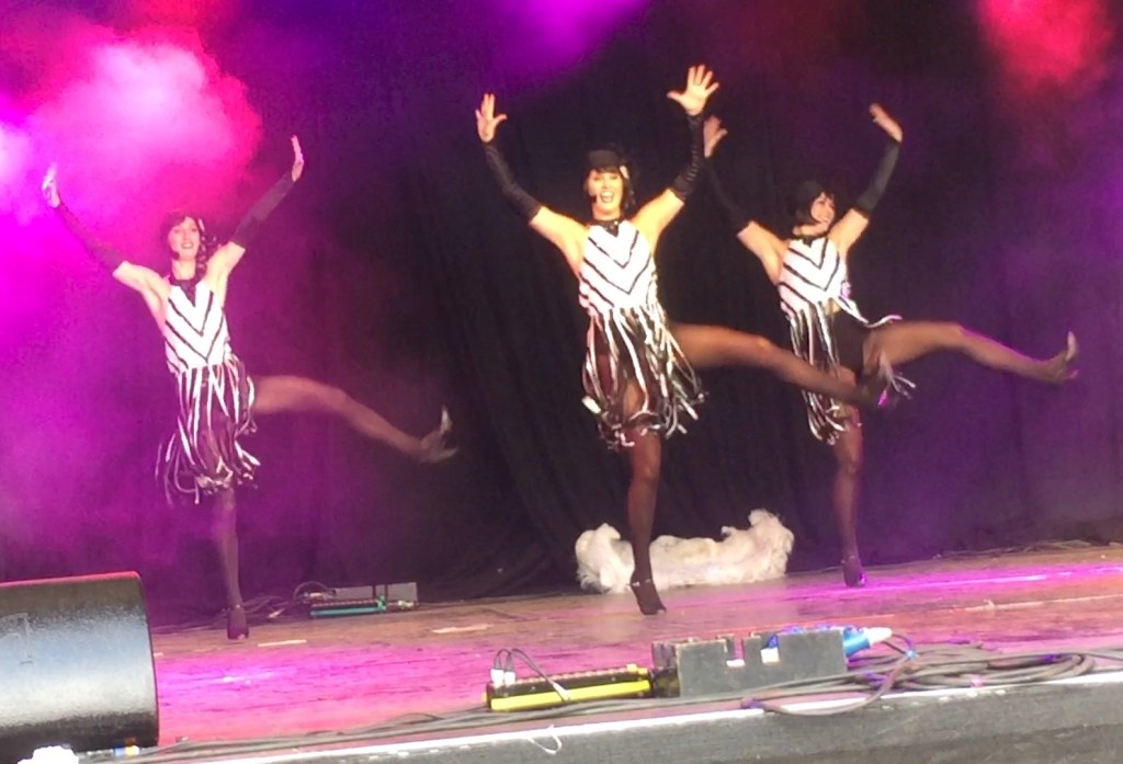 A trio of 1920s style flapper girls dancing on stage, wearing black and white patterned dresses, black stockings, and black sleeves on the forearms.