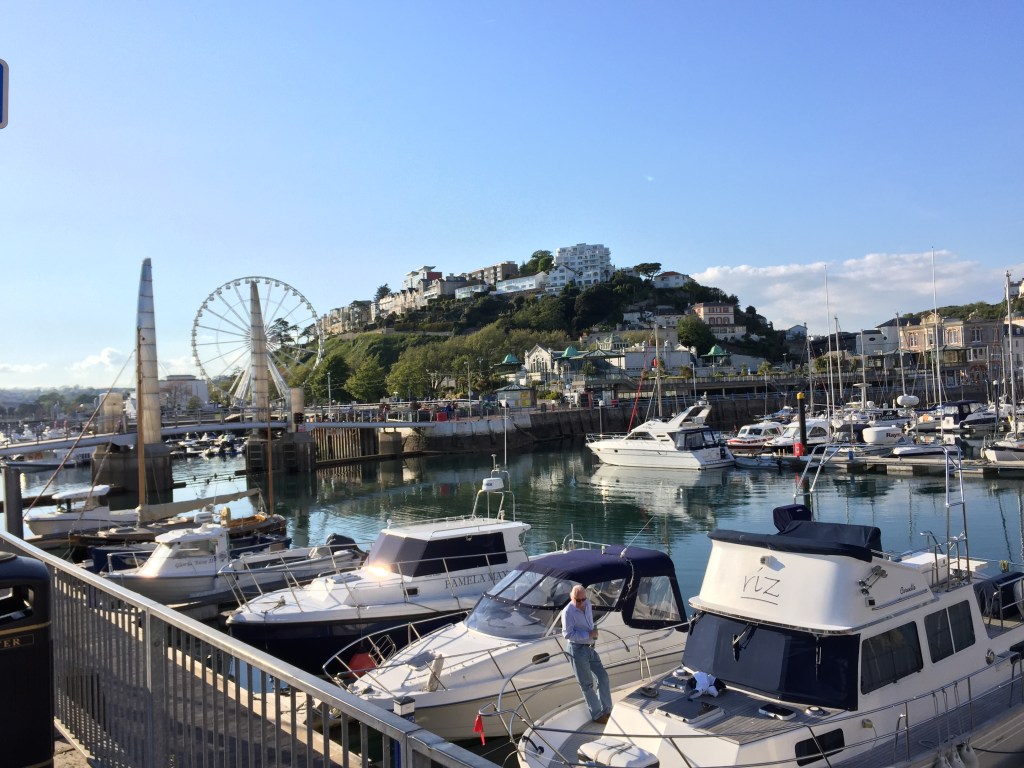 Torquay Harbour in the sunshine. In the background is the Riviera ferris wheel, next to cliffs covered in greenery and buildings.