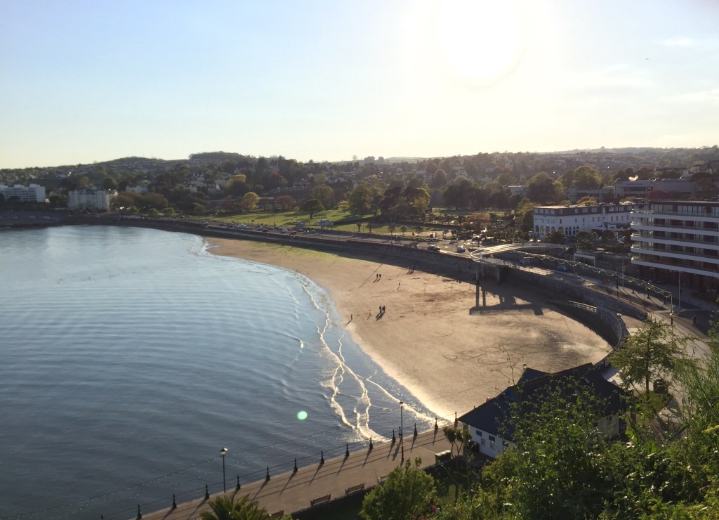 Overhead view of Torquay seafront in the sunshine, from up on the cliffs. The sea is lapping at a long beach stretching into the distance. A long main road runs next to the beach, and on the other side of the road is a large grassy area with some tall trees.
