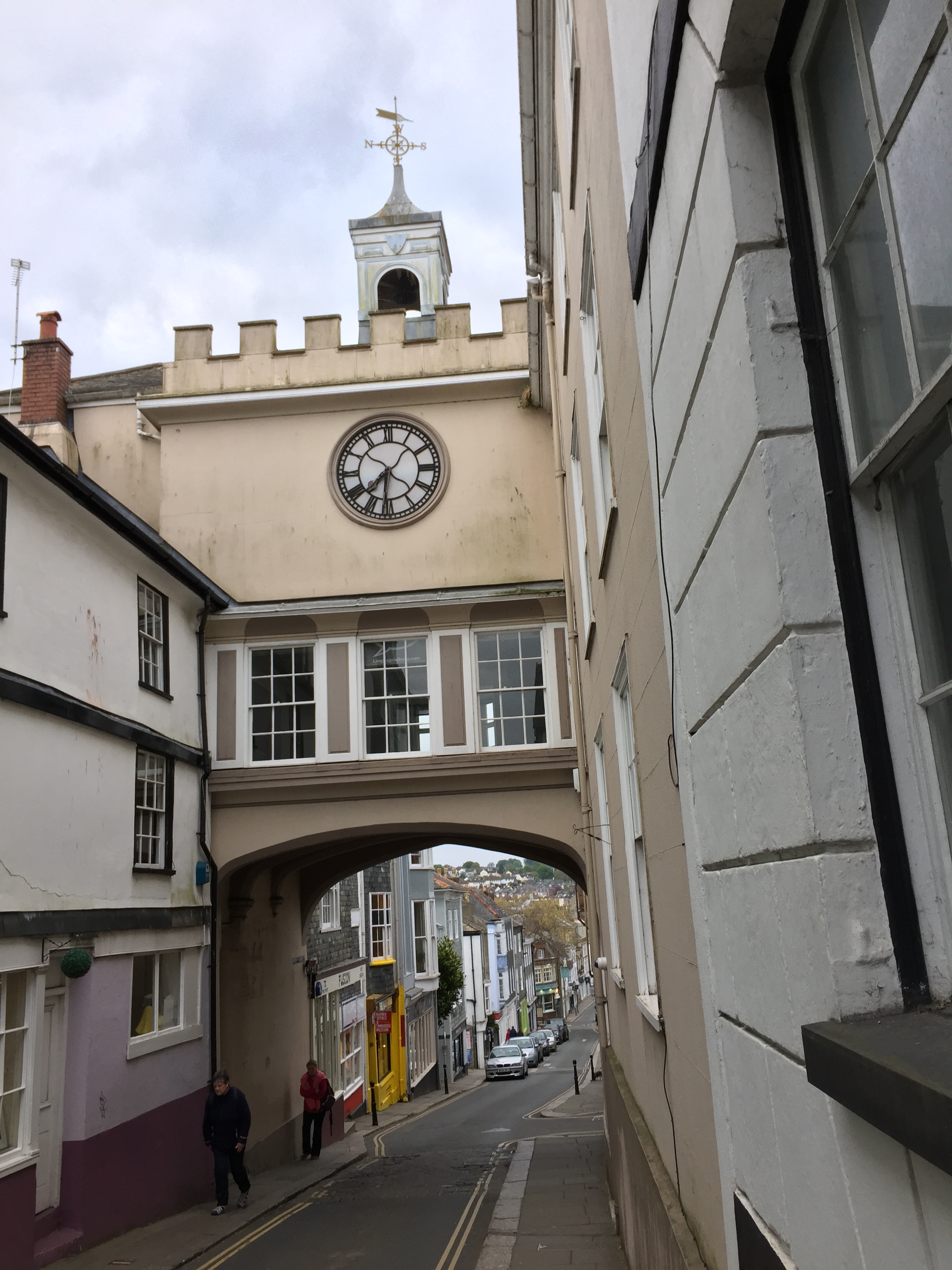A building that bridges over the narrow Totnes high street. The road passes under an archway, above which are three windows, and above those is a white clock face with a black dial and hands. A small tower on top of the structure has a weather vane on it.