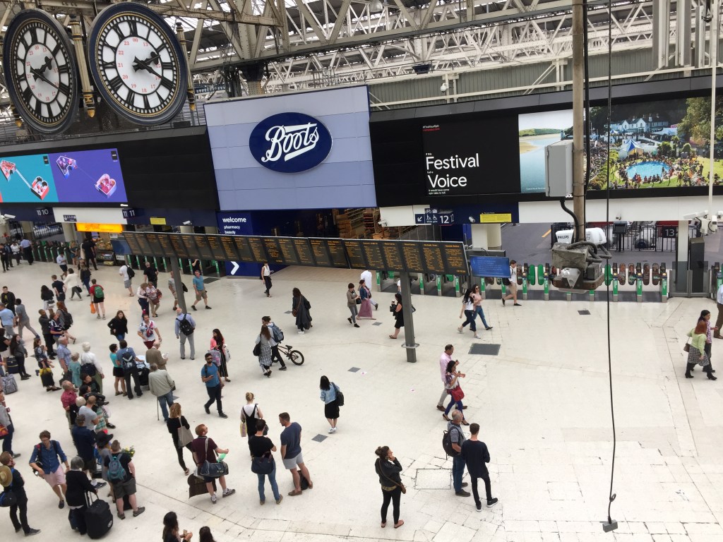 A section of the large, busy concourse at Waterloo Station, including departure boards, a large Boots store, advertising screens and ticket barriers. And at the very top left of the photo is the iconic clock that hangs from the station ceiling, consisting of 4 clock faces arranged in a cube formation, so the time can be read from all sides. Each clock face is white, with black Roman numerals and hands. A circle that touches the bottom of the numerals encloses the centre of the clock face. Within that inner circle are regular numbers, lined up with each of the Roman numerals, that give the PM equivalent numbers in the 24-hour clock - so 13 for 1pm, 14 for 2pm, etc, up to 23 for 11pm and then 00 for midnight.