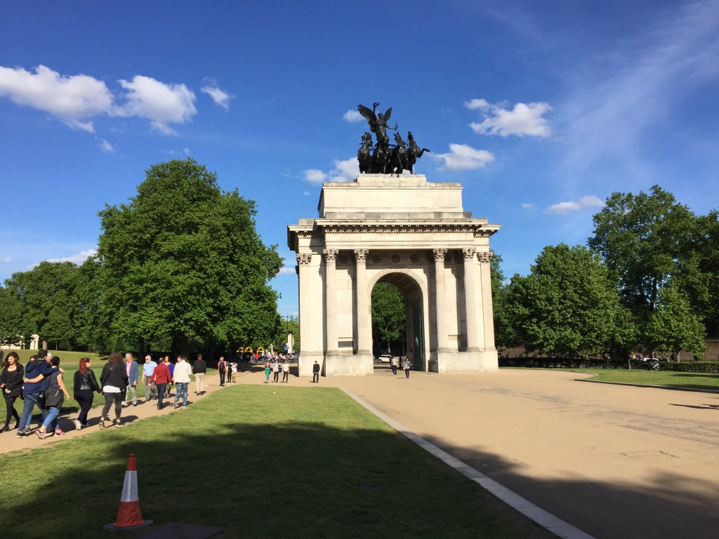 The Wellington Arch on Hyde Park Corner. It is a huge stone archway with ornate cast iron gates, flanked by 2 columns on each side. On top of the arch is a sculpture called the Quadriga of War, depicting 4 horses pulling a chariot, driven by a young boy as a tall female angel with large wings descends from behind.