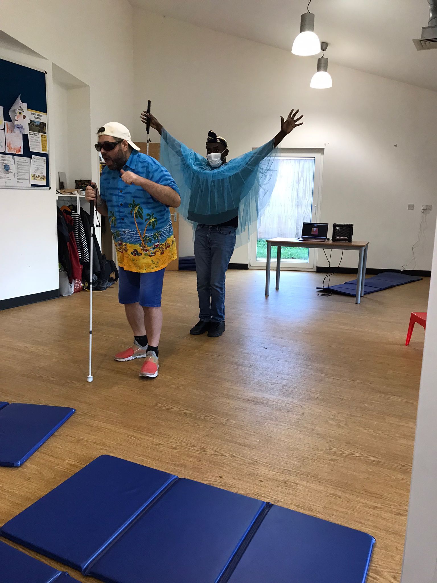 In the Super Power Panto rehearsal studio, the character Billy Bright is wearing a white baseball cap, dark glasses, and a shirt with a colourful seaside cartoon on it. He stands with his mouth wide open as he leans on his long cane. Behind him, The Angel of Light, wearing a veil-like see-through top over a dark t-shirt and jeans, stands with their arms stretched outwards and upwards to each side.