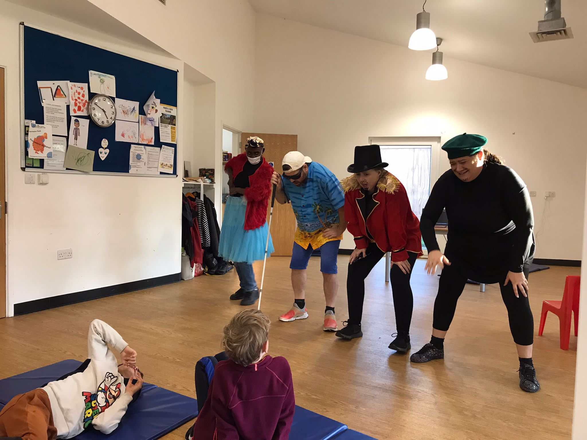 In the rehearsal studio, 4 characters from Super Power Panto stand with their knees bent, as they lean forwards towards the 2 children watching them.