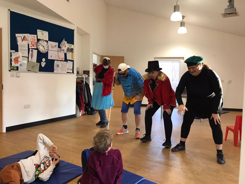 In the rehearsal studio, 4 characters from Super Power Panto stand with their knees bent, as they lean forwards towards the 2 children watching them.