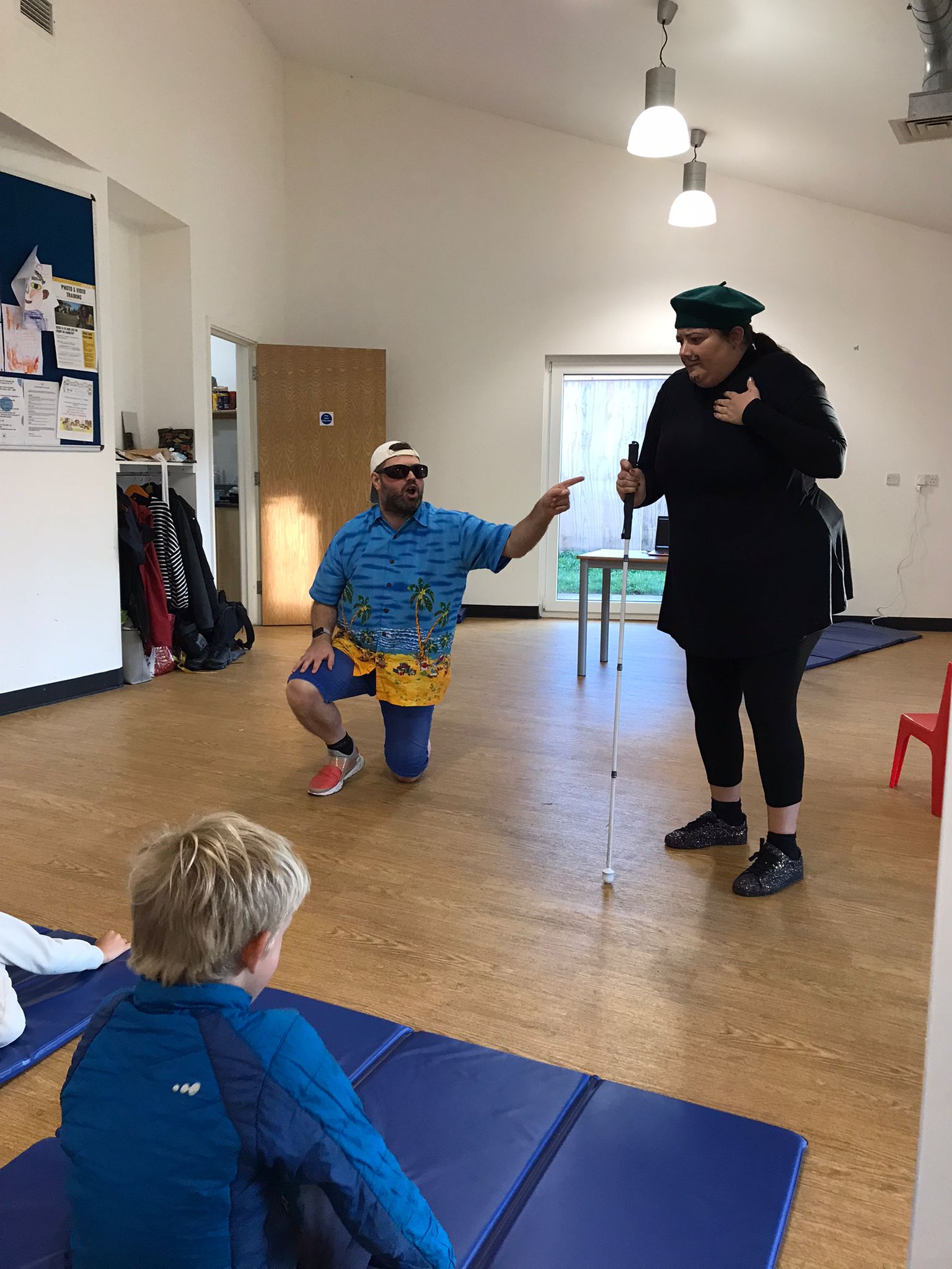 In the Super Power Panto rehearsal studio, the character Billy Bright is down on one knee, as he looks and points towards Claude standing next to him.