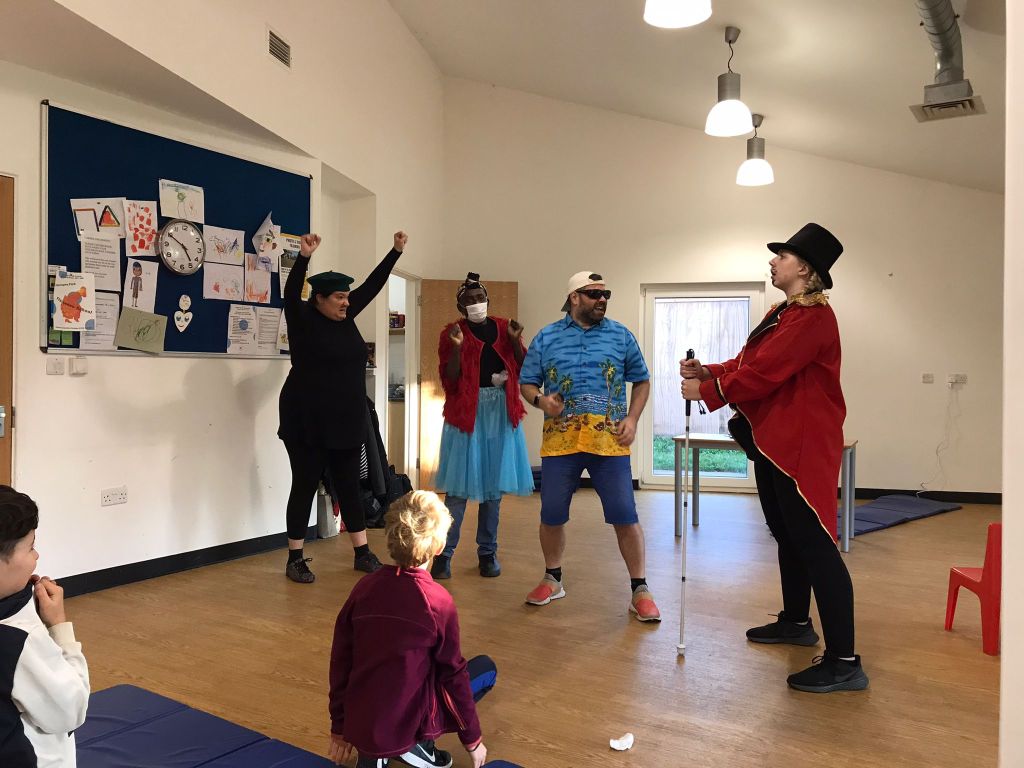 In the Super Power Panto rehearsal studio, a character called Claude holds his fists above his head with an angry look on his face. He and two other characters, Billy and The Angel of Light, are looking towards Sir Sunblock, who stands while gripping a long cane.