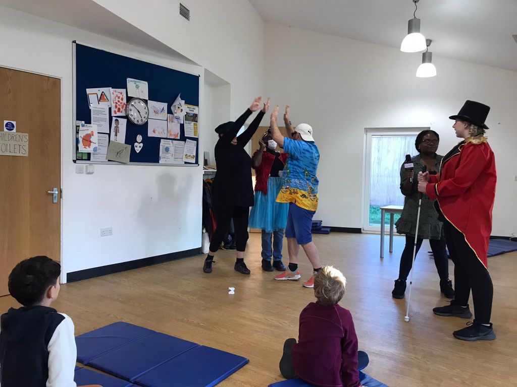 In the rehearsal studio for Super Power Panto, 3 cast members stand in a group high-fiving each other, while the characters called Sunblock and Green Grass talk to each other.