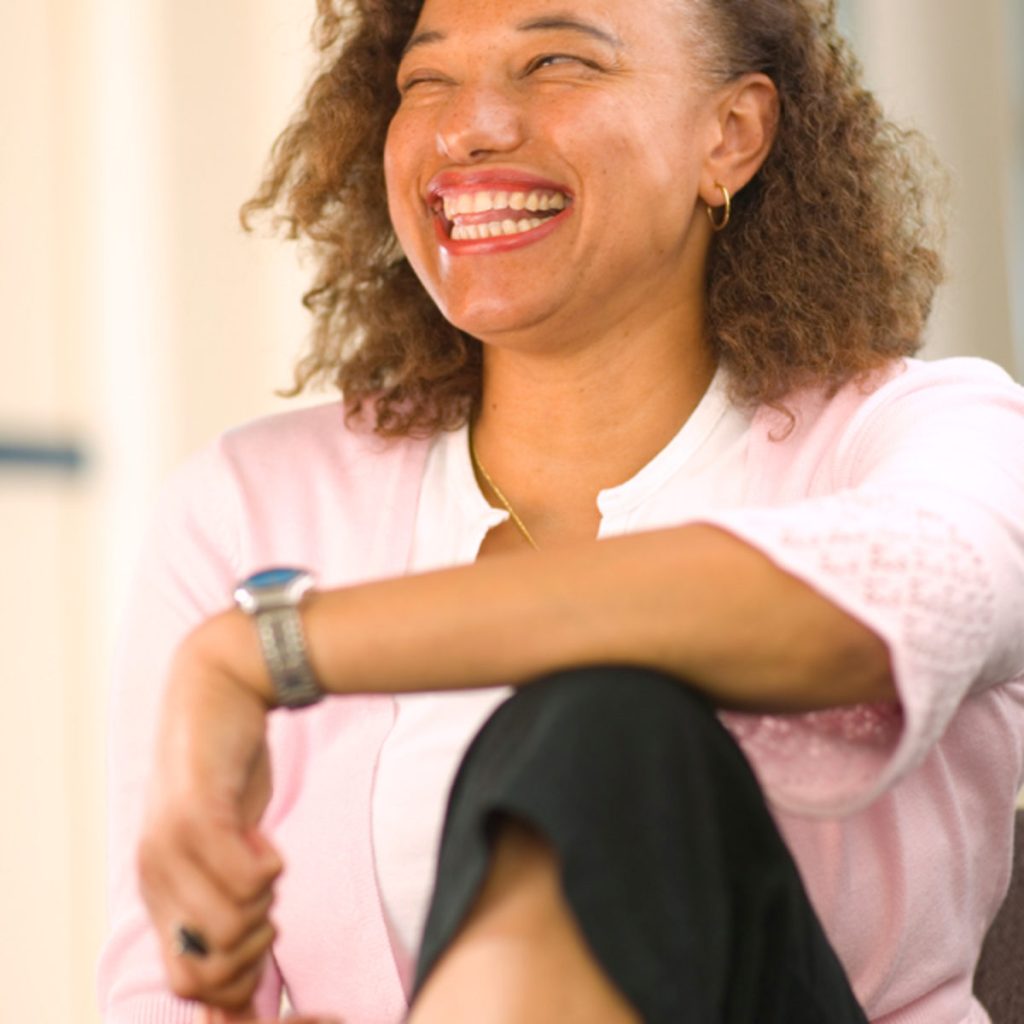 Maria Oshodi laughing gleefully as she sits with her arm resting on one knee in front of her.
