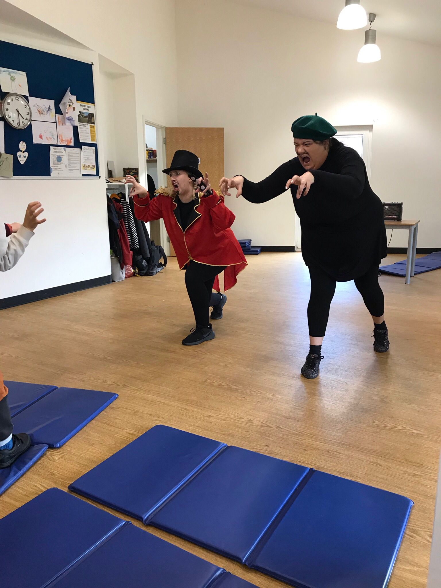 The characters Sir Sunblock and Claude in the Super Power Panto rehearsal room, leaning forward with their hands outstretched in claw shapes. On the left, a child's hand can be seen copying the action.