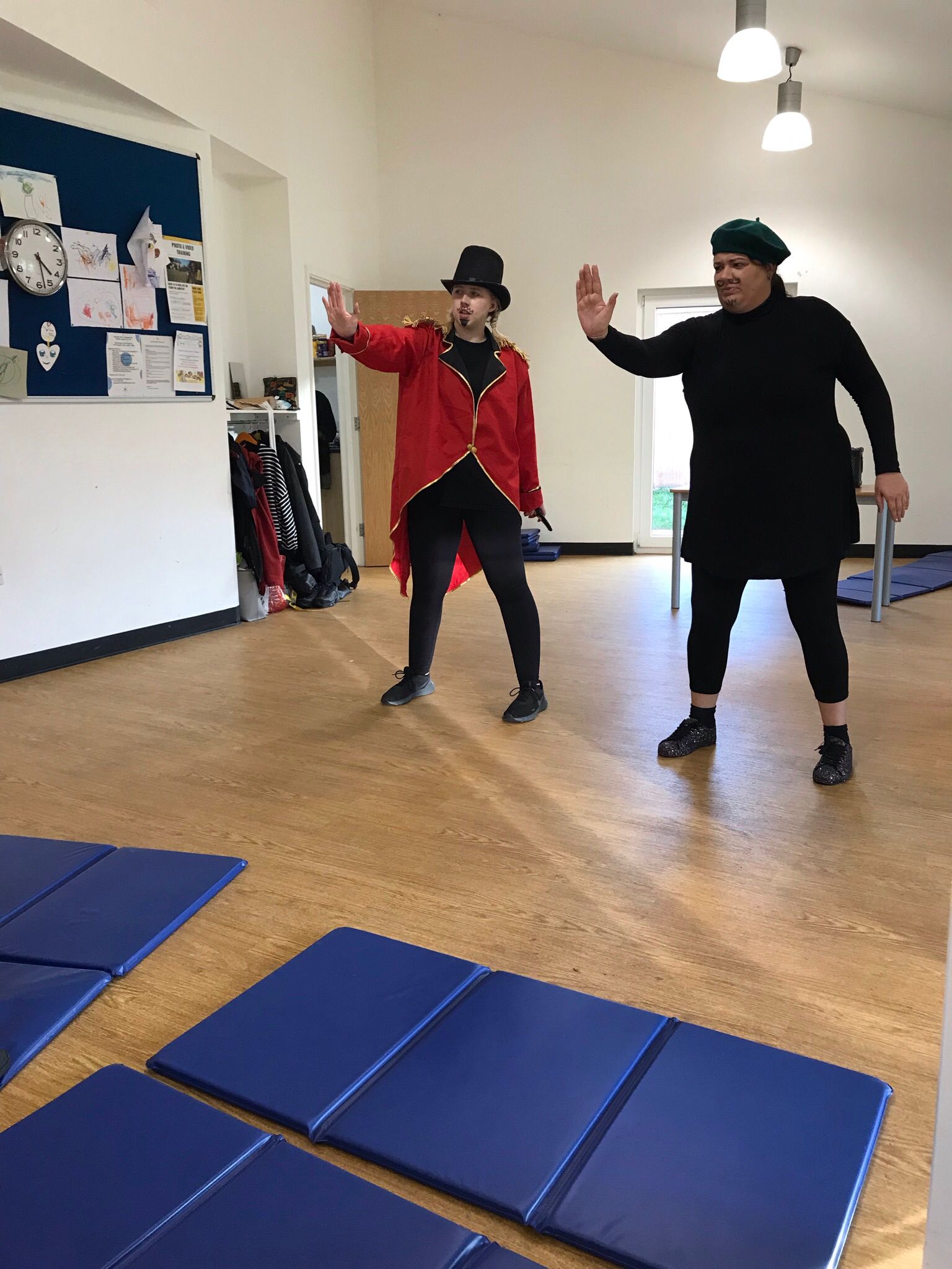 Characters called Sunblock and Claude, in the rehearsal studio for Super Power Panto, standing together, each with their right arm outstretched and the palm of their hand facing away from them, as if signalling to stop or pressing against an invisible wall in front of them.