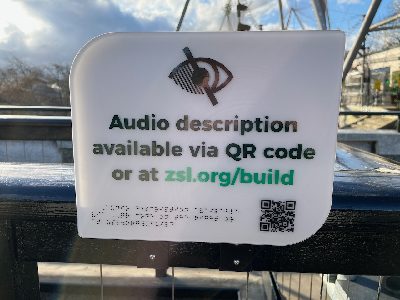 A sign with large black text on a white background that reads Audio description available via QR code or at zsl.org/build. The website address is in green. Above the text is an icon of an eye with a diagonal line across it, such that the left side of the eye is covered in lines while the right side is unobscured. Below the text are 3 lines of braille and a QR code.
