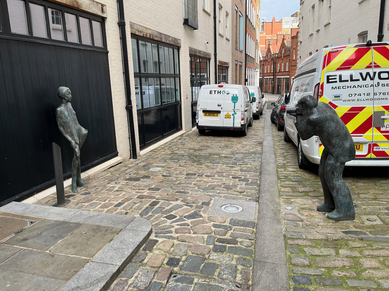 Bronze statues representing Terence Donovan, standing in the middle of a cobbled street, photographing Twiggy, posing in front of a black wall.