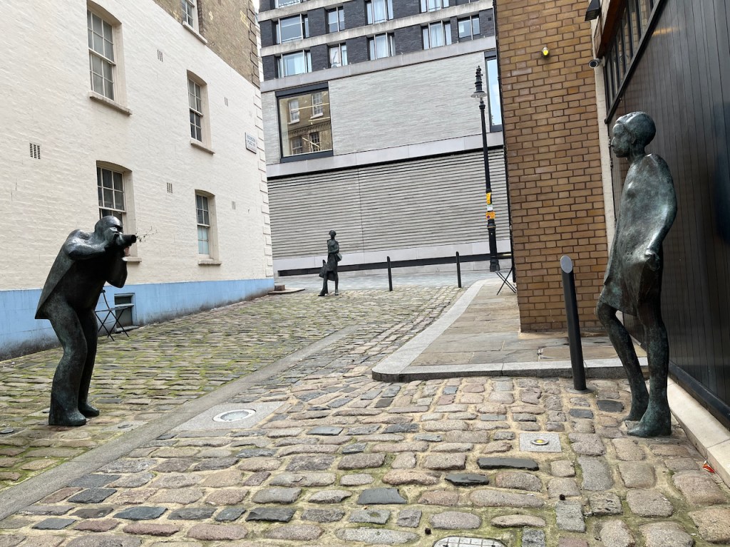 Bronze sculptures of Terence Donovan photographing Twiggy in a cobbled street, with a further statue of a female shopper stopping to look at them at the far end of the street.