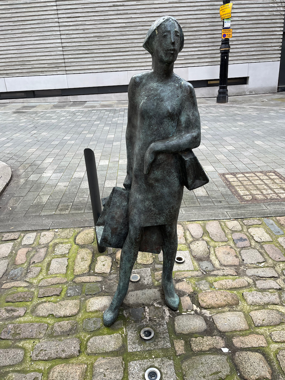Bronze sculpture of a lady carrying a large bag in her right hand, and a small handbag hung over her left forearm.