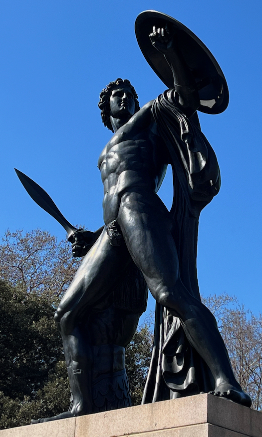 An 18 foot bronze statue of the Greek hero Achilles, standing with legs astride as he looks to his left and holds up a large round shield, while in his right hand behind him he holds a sword.