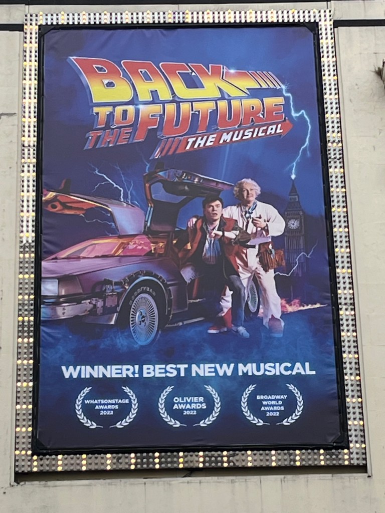 Poster for Back To The Future The Musical, showing Marty and Doc Brown posing beside the DeLorean car, which has its gull-wing style doors opened upwards and outwards.