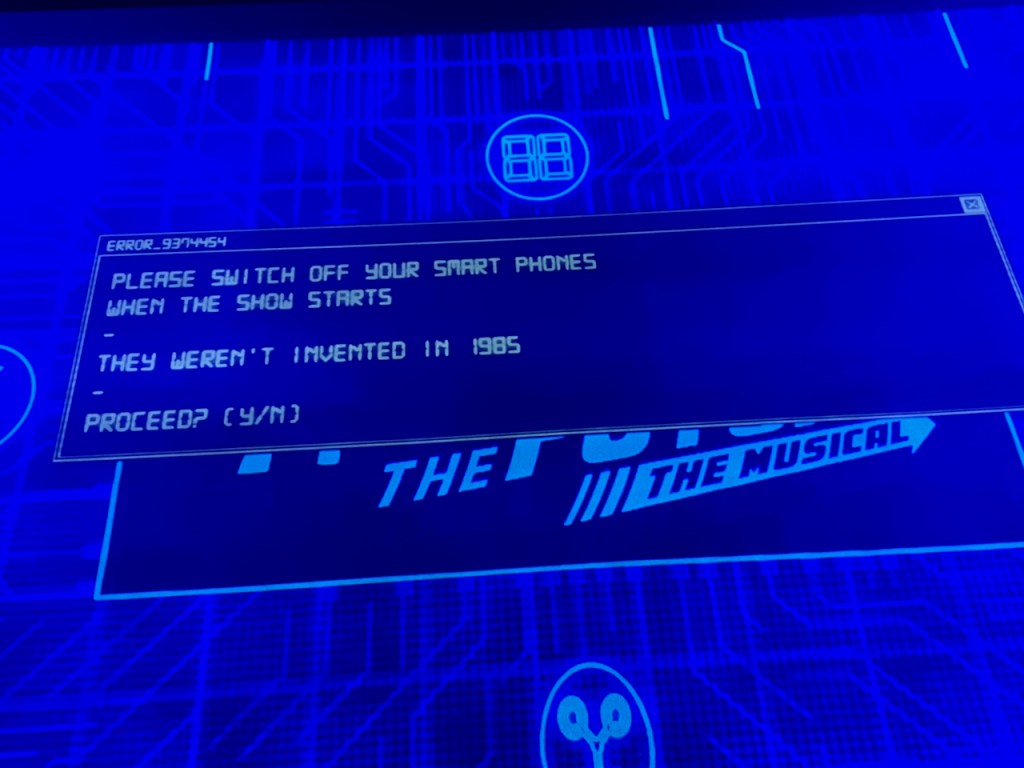 A message on the projection screen at the front of the stage for Back To The Future The Musical. It reads Error 9374454. Please switch off your smart phones when the show starts. They weren't invented in 1985. Proceed? Y or N.