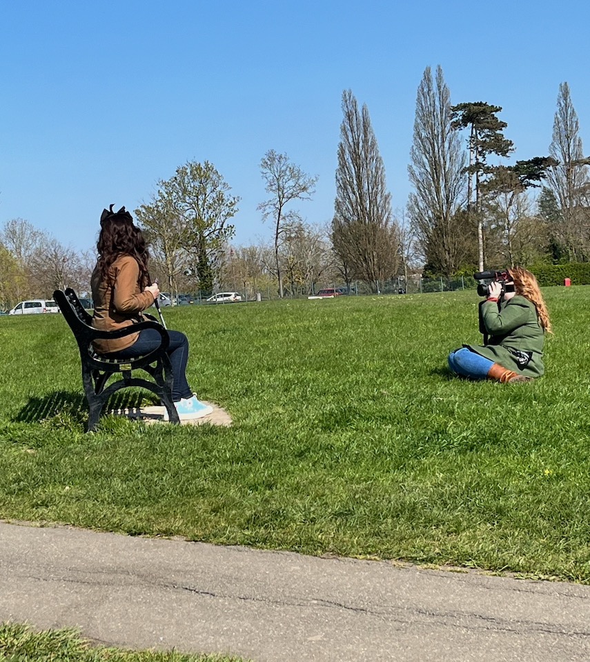 Taylor sitting on a bench in a grassy area of Danson Park, holding her long white cane, while Emily sits on the grass and films her.