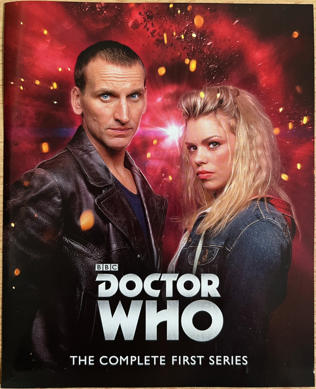 The front side of the flyer included in the Doctor Who Series 1 Blu-ray set, with an image of Christopher Eccleston's Doctor and Rose Tyler standing together, against a background of red clouds and stars in space.