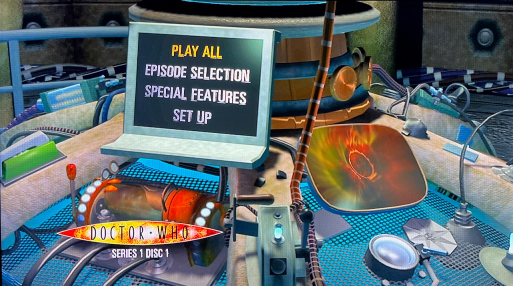Blu-ray menu for Doctor Who Series 1. The 4 options - Play All, Episode Selection, Special Features and Set Up - are displayed on a computer screen on the Tardis console, with various lights, levers and buttons around it, and another screen to the right with a view of the swirling time vortex.