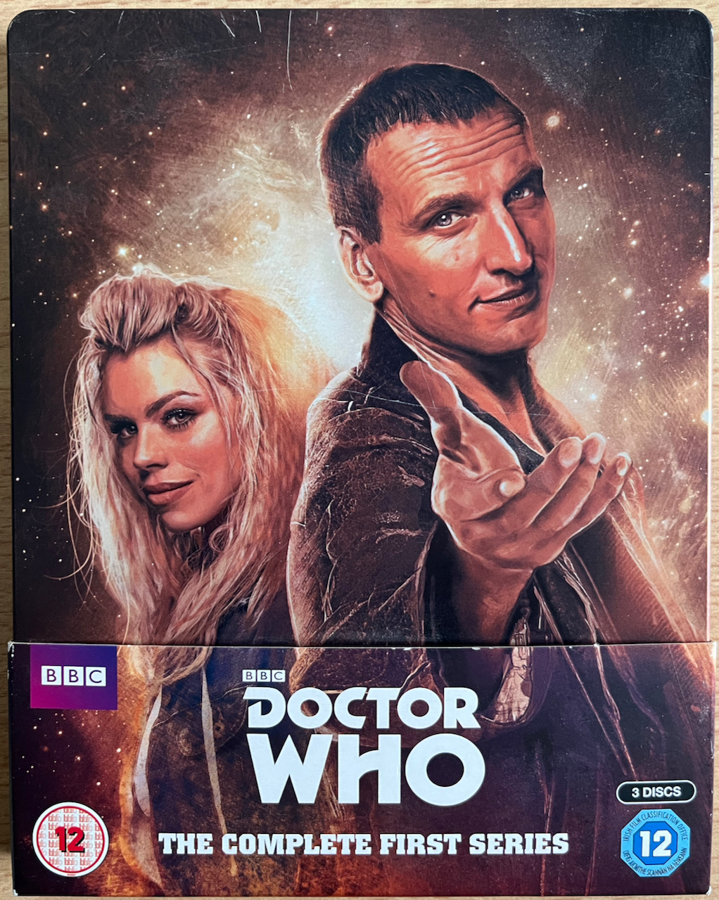 The front of the Blu-ray steelbook for Doctor Who Series 1, showing artwork of Christopher Eccleston's Doctor reaching out to us with his hand, as Rose Tyler stands behind him, also looking at us.