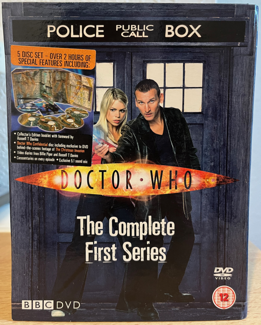 Front of the Tardis-shaped DVD box set for Doctor Who, The Complete First Series. Christopher Eccleston's Doctor and Rose are stood in front of the Tardis doors, with the Doctor Who logo across their chests, and a sticker to the left of Rose details the contents of the set, stating there are 5 discs with over 2 hours of special features.