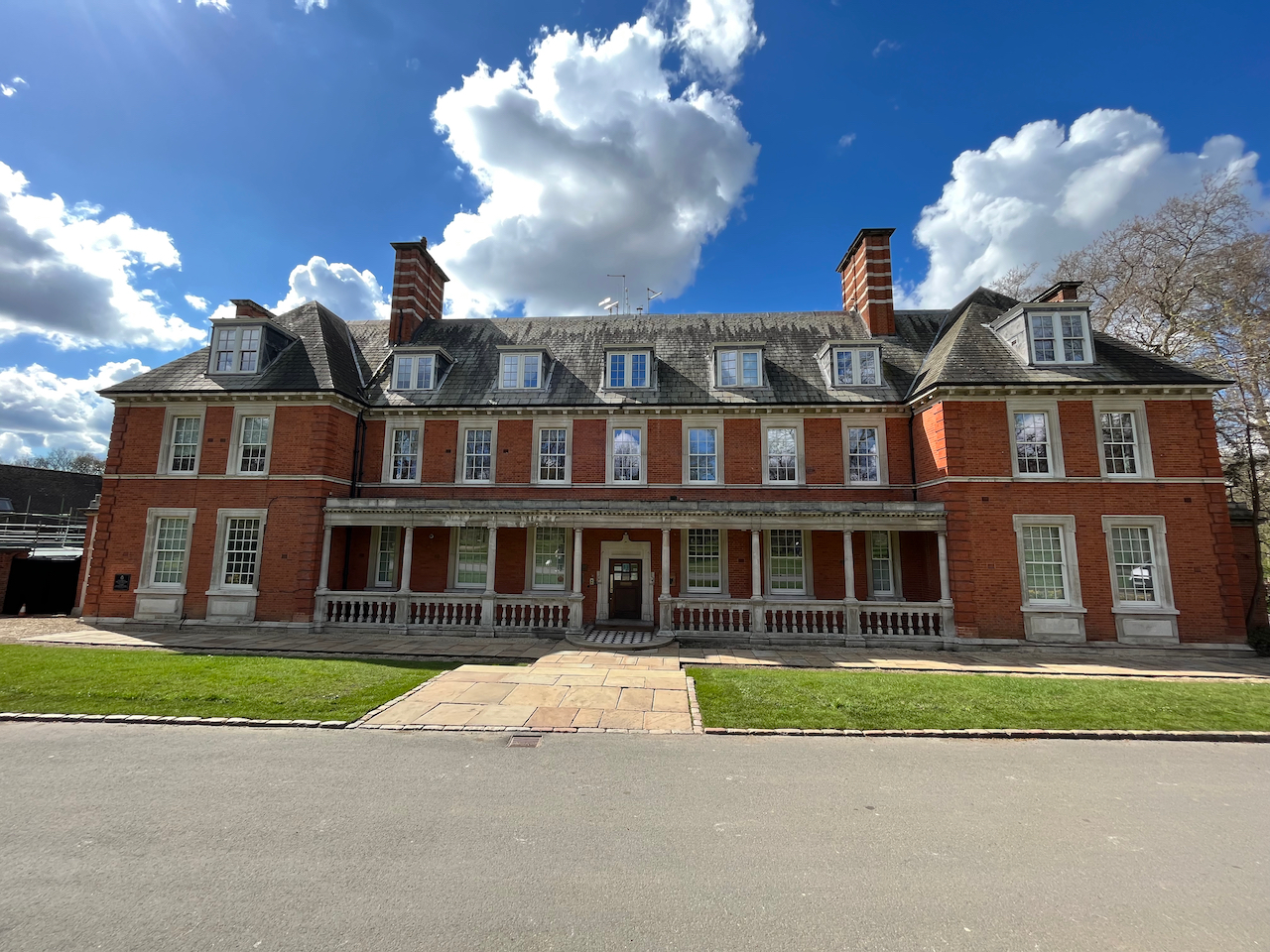 The Old Police House, a very wide red-brick 3-storey building, with the top floor being within the sloping roof. The sections of the house at either end have 2 windows on the first 2 floors, then a slightly smaller window in the centre of the roof above. In the central section, the ground floor has a pathway leading to the front door, with 3 windows on each side of the doorway. A canopy over the ground floor is supported by pillars connected to stone railings on the floor. Above, there are 7 windows on the first floor, and 5 in the roof space, with a wide chimney stack at each end.
