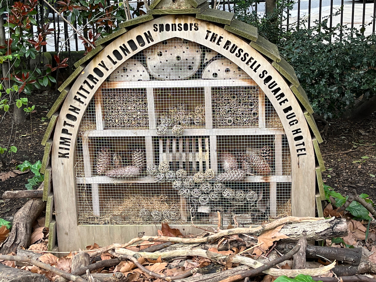 An arch shaped wooden shelter for bugs, with text curving around the top at the front that reads Kimpton Fitzroy London sponsors The Russell Square Bug Hotel. Inside the arch are various compartments containing small items suitable for different types of insects.