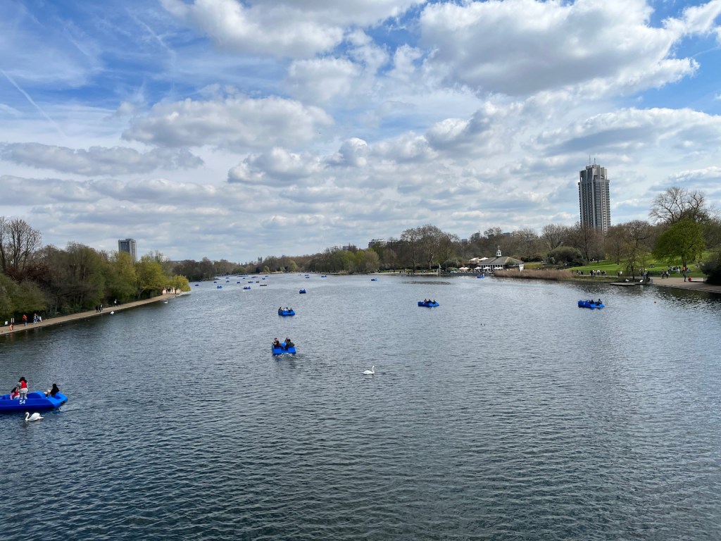A view down the very long and wide Serpentine Lake in Hyde Park, including a few people sailing in blue pedal boats, and trees along each bank.