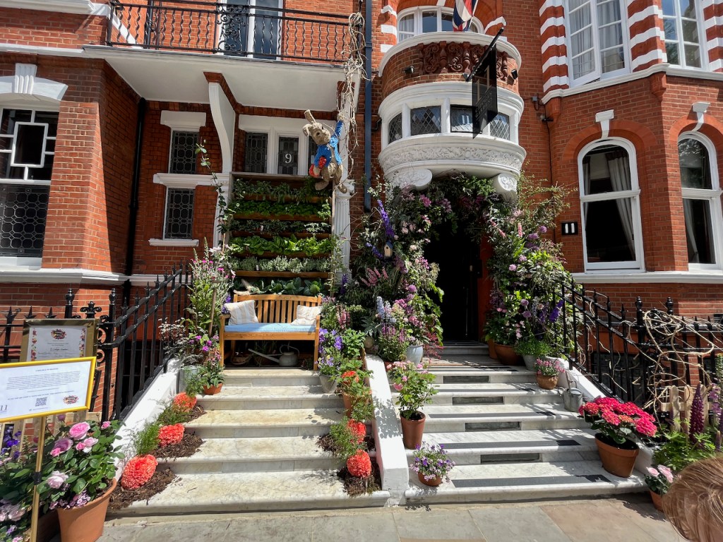 A floral display representing Peter Rabbit, across the entrances of 9 & 11 Cadogan Gardens. On the left, Peter Rabbit clings on to some branches as he dangles above a wooden bench on the top of the steps leading up to the house. Behind the bench, in the doorway, is shelving crammed full of green plants. The entrance on the right, meanwhile, has colourful flowers in an arch around the doorway. And on the 5 steps leading up to both houses, there are small plant pots containing colourful flowers on the left and right side of each step.