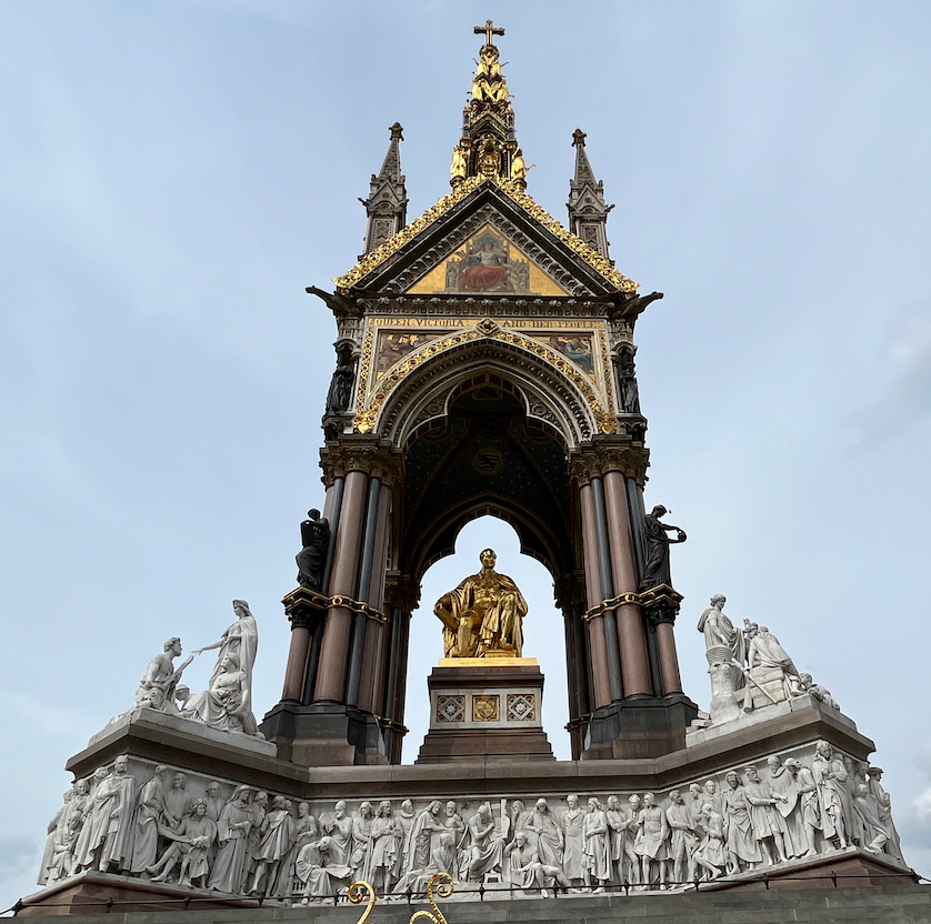The Albert Memorial, a very tall, highly ornate, gothic style tower, sheltering a gilt statue of Prince Albert seated on an altar. The tower stands on a wide stone plinth, on each end of which are groups of stone statues, while a stone frieze of sculptured figures runs across the bottom of the plinth.