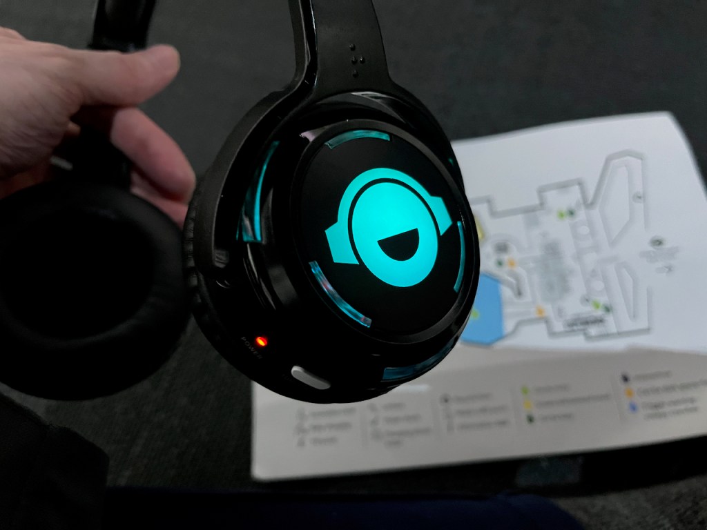 The audio description headset for the BBC Earth Experience, with a bright coloured symbol representing a smiling face wearing headphones on the outside of the padded earpiece. The symbol is lit up in blue, and the colour is changed by pressing a white button on the underside of the earpiece to switch between the audio description channels.