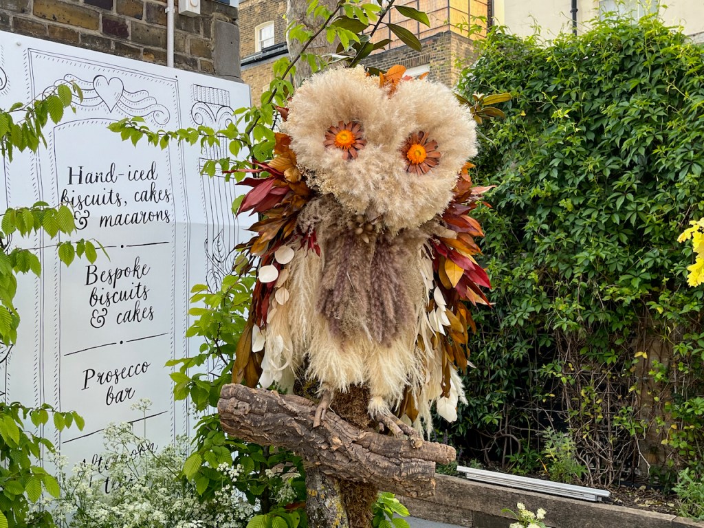 A model of an owl with flowers for eyes standing on a thick branch in front of some greenery. A sign next to it says the shop, called Biscuiteers, does hand-iced biscuits, cakes and macarons, bespoke biscuits and cakes, and has a Prosecco bar.