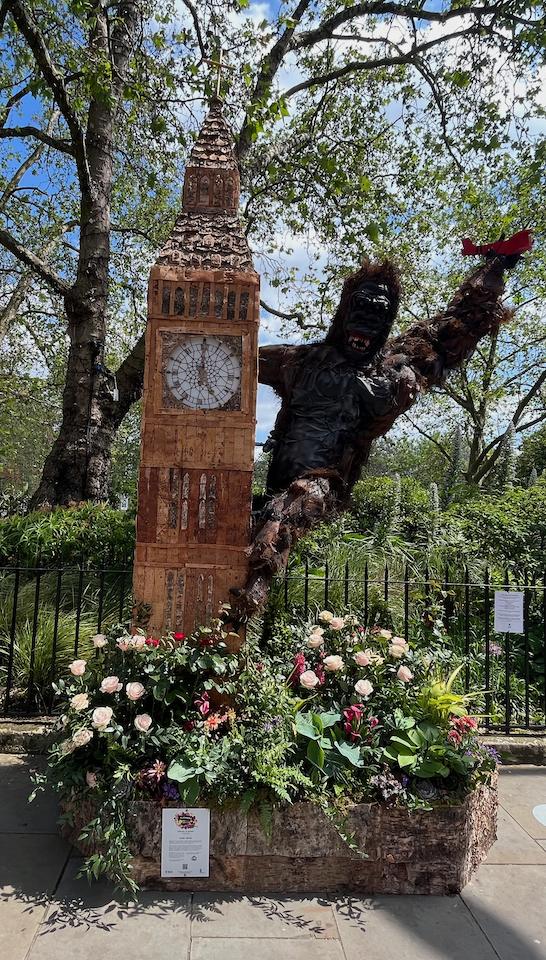 A large model of the black gorilla King Kong clinging on to Big Ben with its legs and right arm, while in its left arm it holds a small bi-plane that it has caught flying past. The whole model stands in a small bed of colourful flowers and greenery.