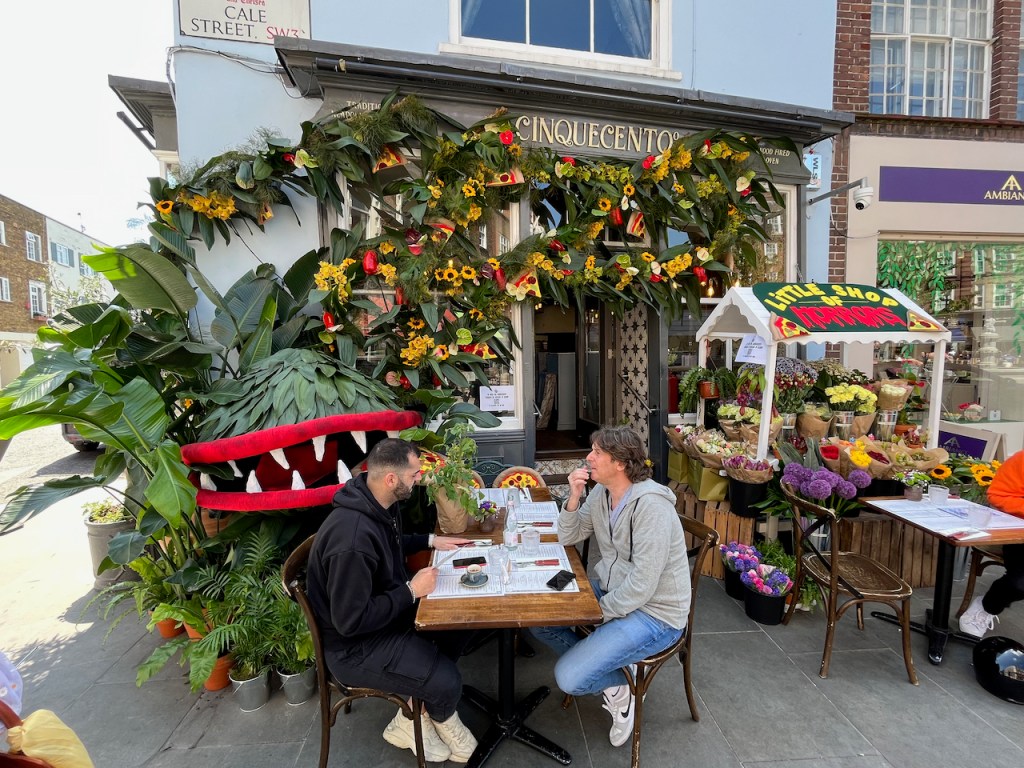 Floral display representing The Little Shop Of Horrors, with a large round green plant that has opened its red lips to reveal sharp teeth in its wide mouth. The rest of the display is mainly made up of yellow flowers and greenery stretched over the front of the restaurant, but there is also a small market stall with the wording Little Shop Of Horrors on its roof, that has lots of colourful flowers inside it.
