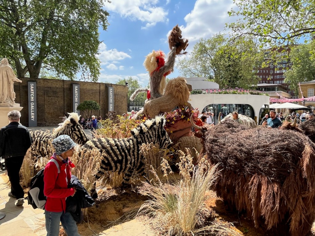 Side view of a floral display representing The Lion King, with Rafiki the mandrill holding up Simba the baby lion, while standing on a large rock next to an adult lion. A couple of large zebras are also standing near the rock.